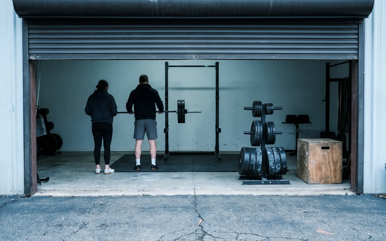 Two athletes seen from behind set up a barbell in a squat rack inside an open garage, a plate tree and a wooden plyo box beside them.