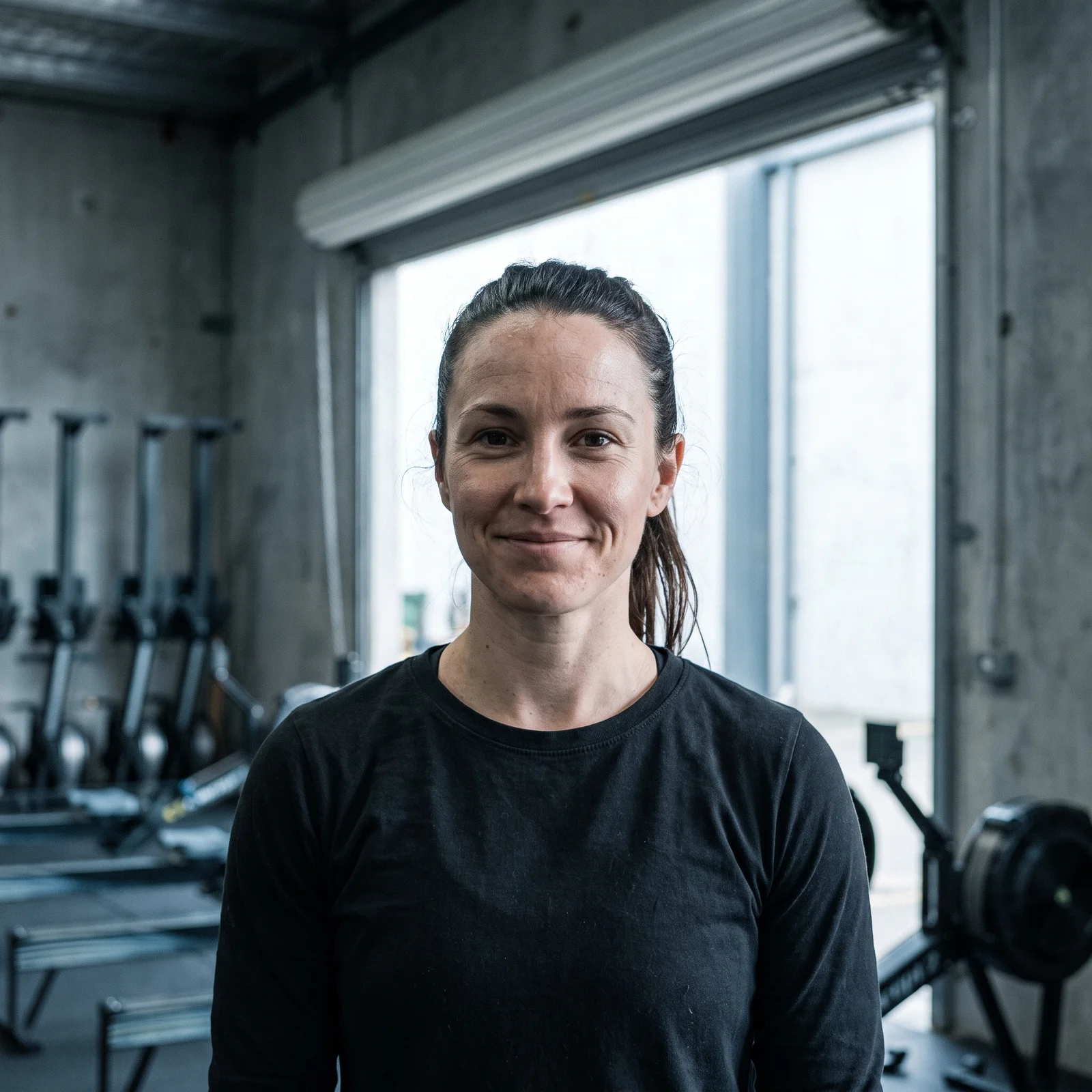 Headshot of Renata, a coach with her hair tied back in a black long-sleeve shirt, rowing machines behind her.